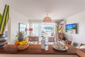 A modern living room with a wooden coffee table, a fruit bowl, and a bottle of liquor. There is a view of the beach through the window.