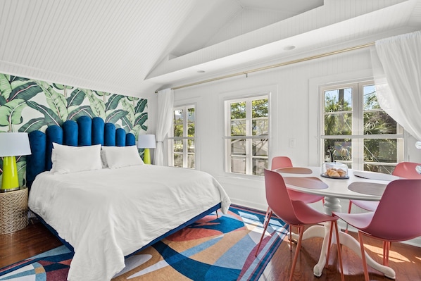 Una habitación de hotel moderna con una cama grande, una zona de comedor con sillas rosas y vistas al verdor por las ventanas.