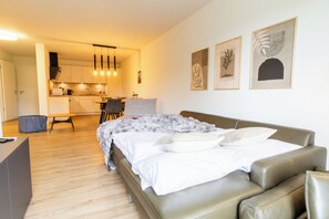 A modern bedroom with a bed, a desk, and a kitchenette in the background.