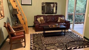 A living room with a brown leather couch, a wooden chair, a black coffee table, a patterned rug, a wooden ladder, and a large window with a view of greenery. - The Pecan at Hamilton Cove ~ Cabin Twenty ~ Lakefront ~ Pet Friendly (Royal)
