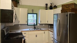 A compact kitchen with white cabinets, a stainless steel refrigerator, and a window with a view of greenery. - The Pecan at Hamilton Cove ~ Cabin Twenty ~ Lakefront ~ Pet Friendly (Royal)