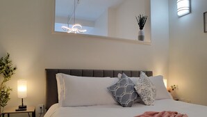 Una cama bien hecha con ropa blanca y almohadones decorativos, una mesita de noche con lámpara y una pequeña planta en maceta.