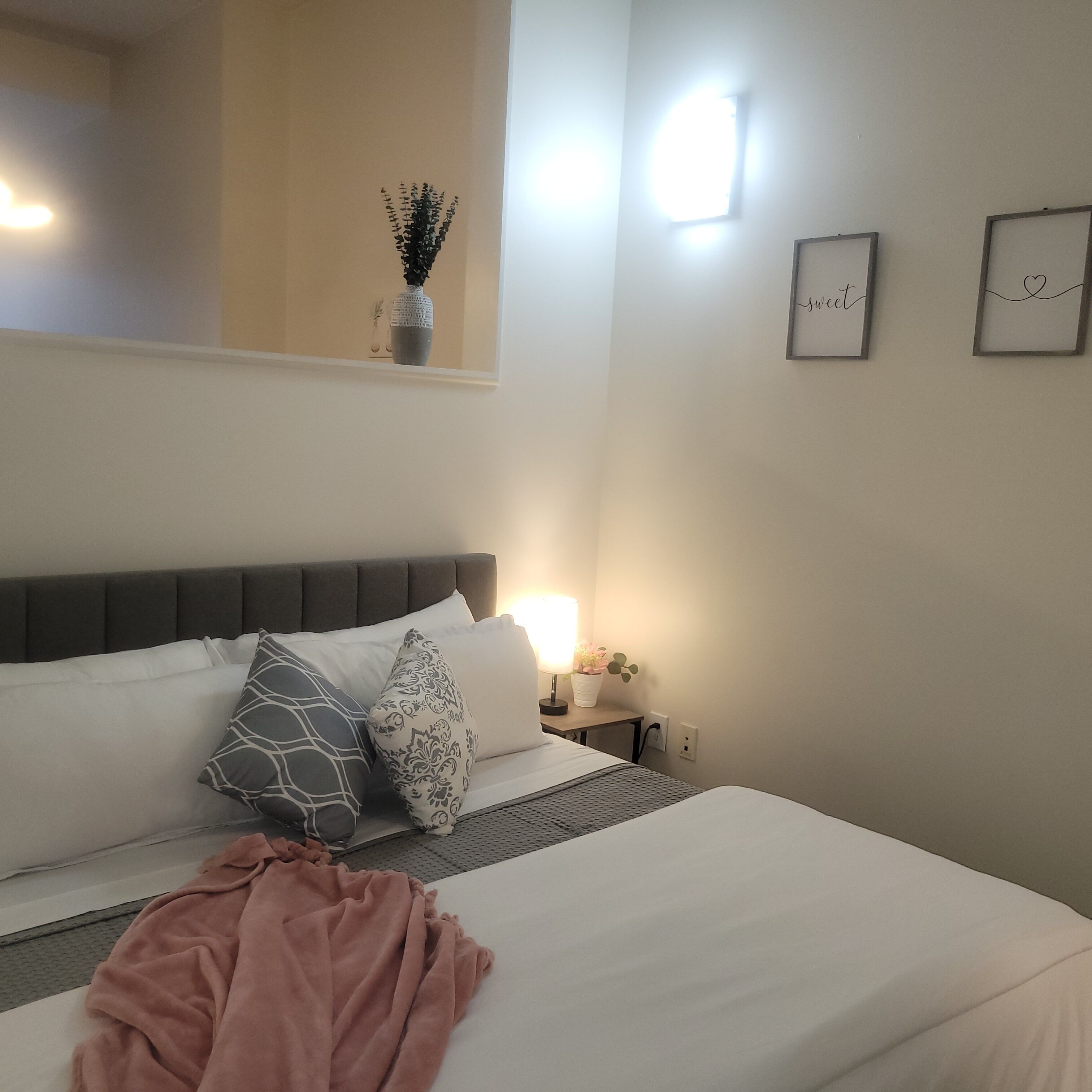 Een slaapkamer met een bed, kussens, een nachtkastje met een lamp, een klein plankje met een plant en een kunstwerk aan de muur.