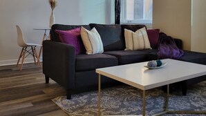Un salón moderno con un sofá gris oscuro, una mesa de centro blanca y una alfombra estampada. Hay una ventana con persianas, un equipo de aire acondicionado blanco y dos cuadros abstractos en la pared.