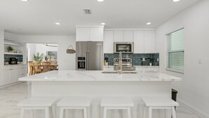 A modern kitchen with white cabinetry, a large island with bar stools, stainless steel appliances, and a dining area with wooden chairs.