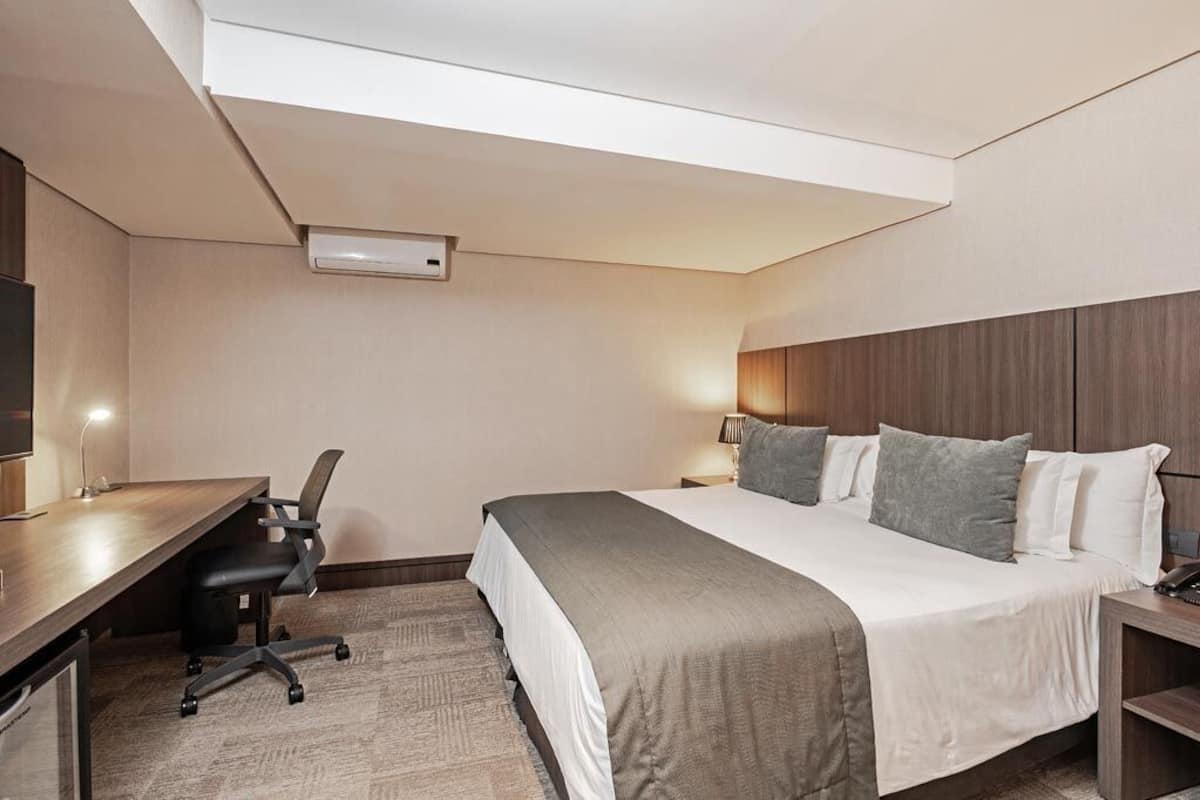 A hotel room with a bed, bedside table, lamp, desk with chair, and an air conditioning unit.