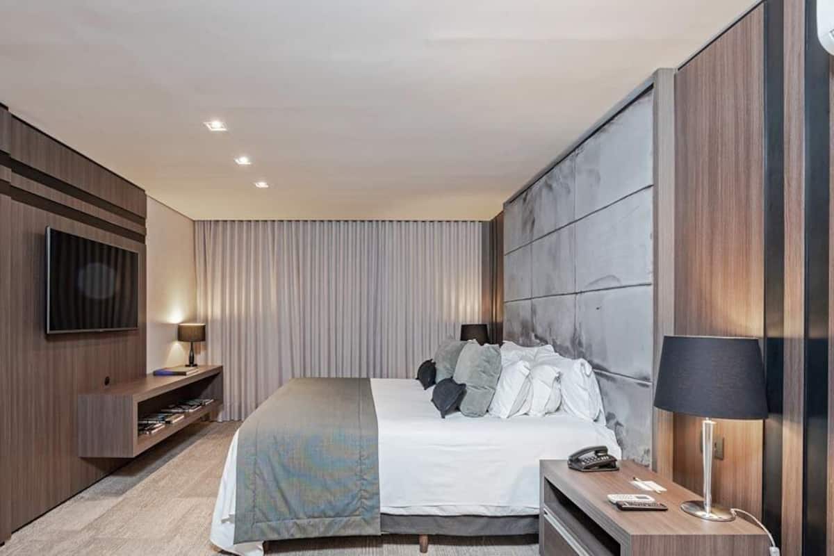A modern hotel room with a large bed, a flat-screen TV mounted on the wall, a bedside table with a lamp, and a built-in shelf with books.