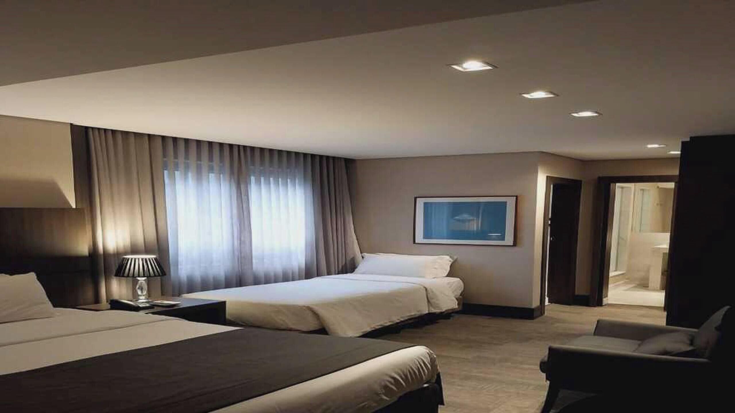 A hotel room with two beds, a desk, a chair, a lamp, and a painting on the wall.
