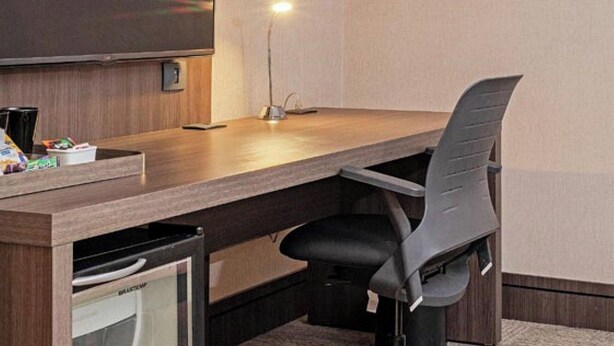 A hotel room with a flat-screen TV mounted on the wall, a desk with a chair, and a desk lamp.