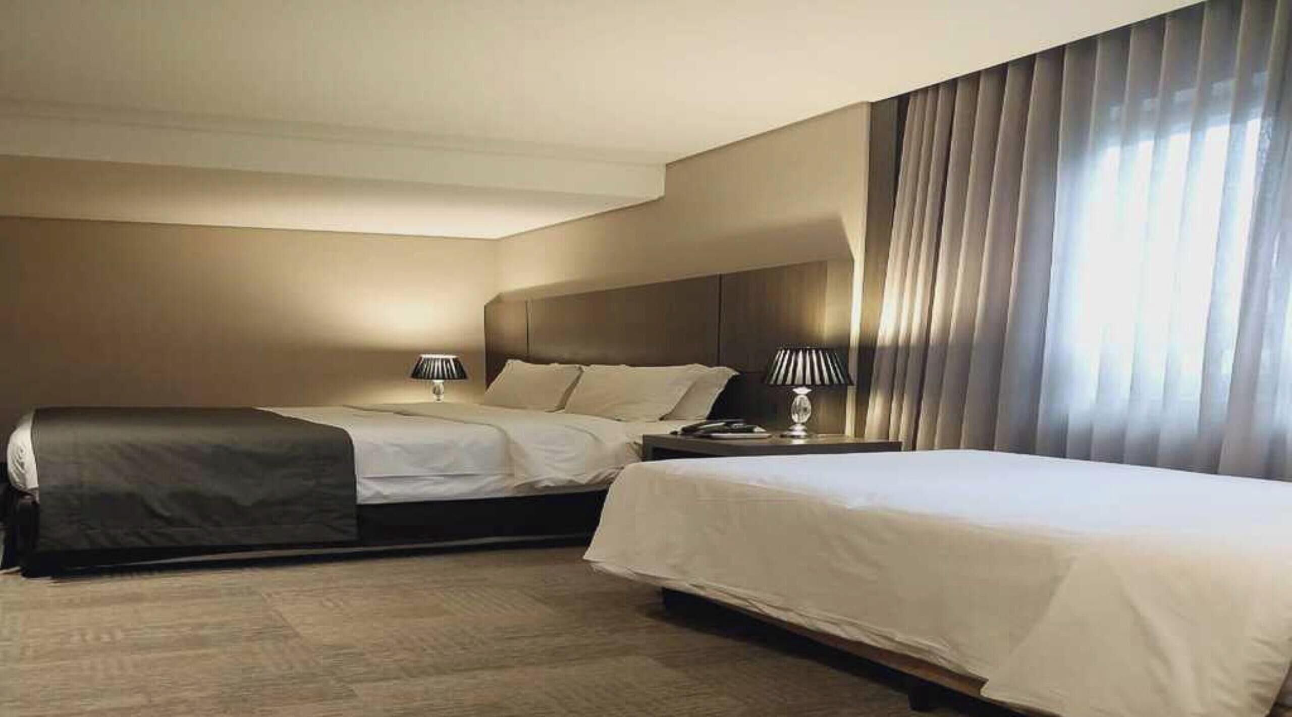 Hotellihuone, jossa on kaksi sänkyä, yöpöytä, lamppu ja suuri ikkuna, jossa on verhot.