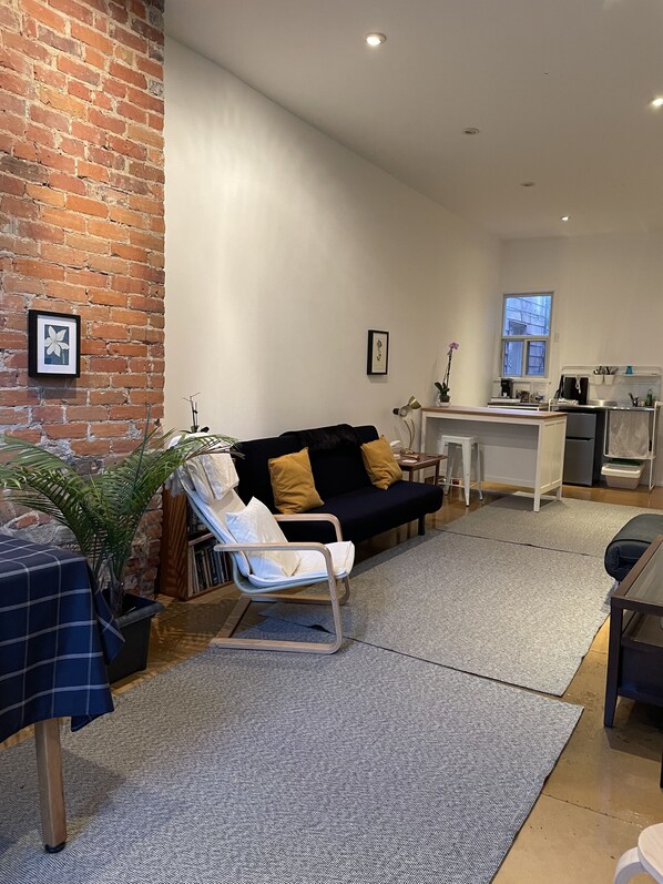 A living room with a black sofa, a white chair, a coffee table, a brick wall, and a window. - Modern Ossington Apartment (Toronto)