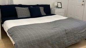 Un lit bien fait, avec une couette à carreaux gris, une tête de lit bleu foncé et une table de chevet blanche ornée d’une lampe et d’un tableau encadré.