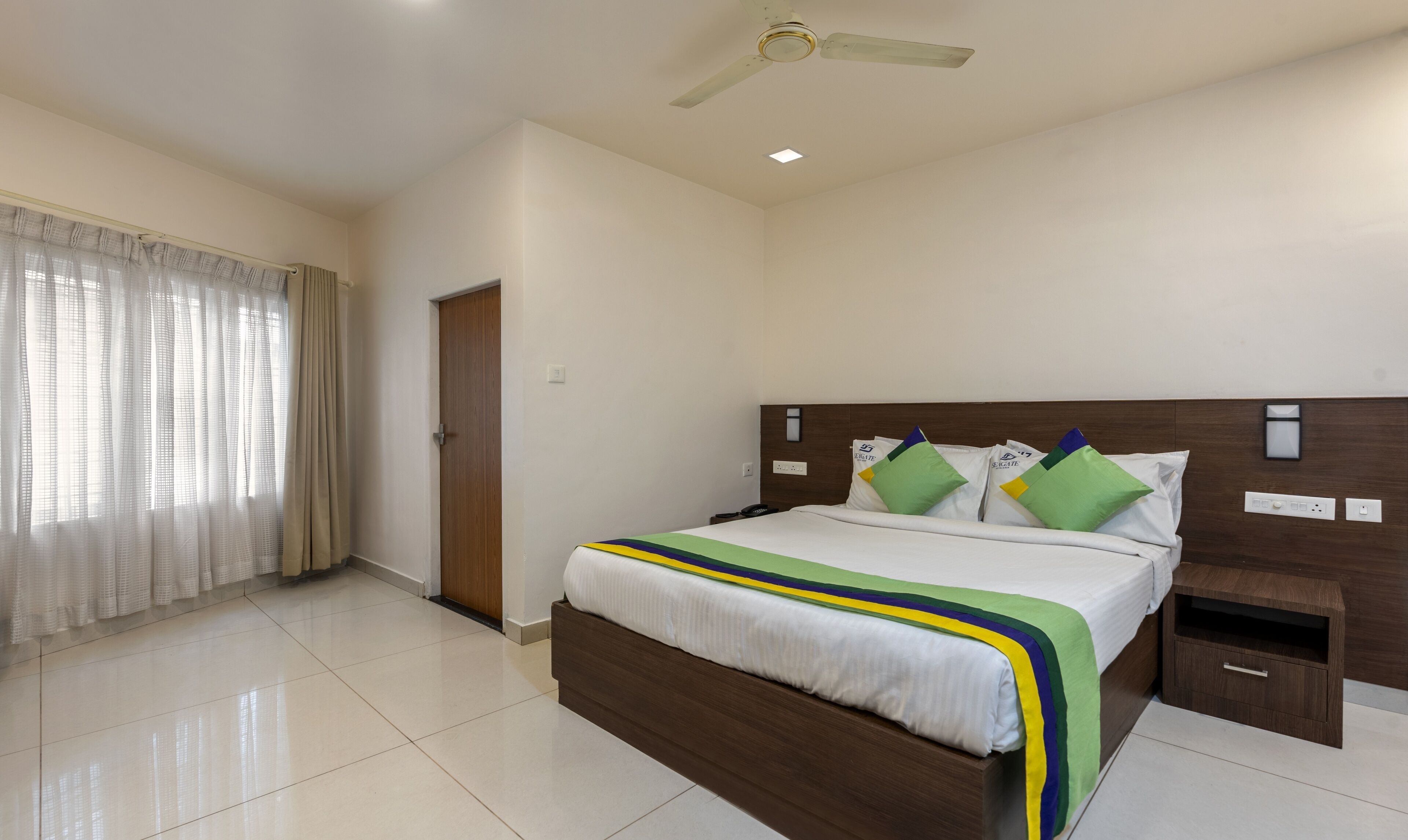 A hotel room with a bed, a nightstand, a ceiling fan, and a window with curtains.