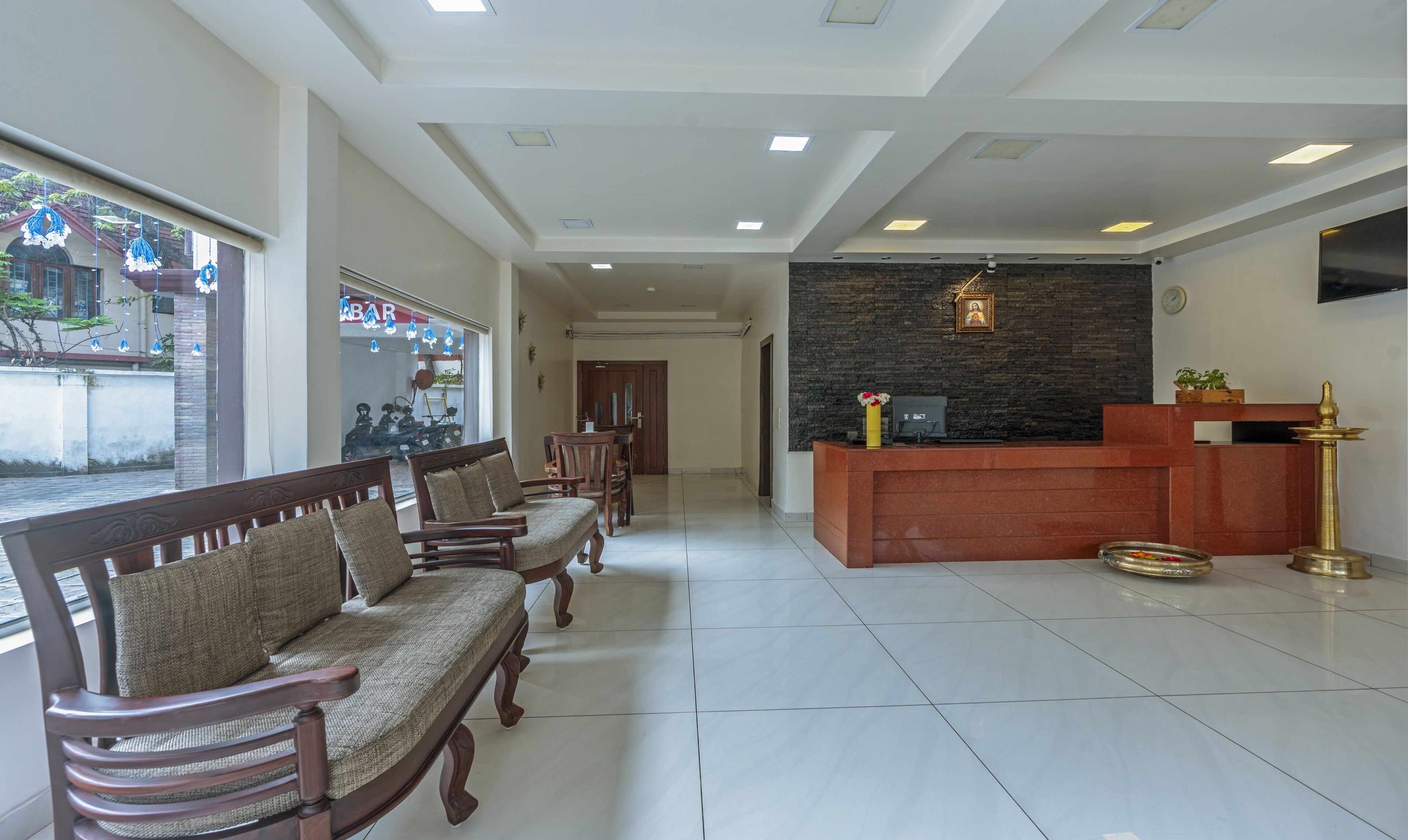 A reception area with a reception desk, a wall-mounted TV, and a reception counter. There are wooden chairs and a sofa arranged for guests.