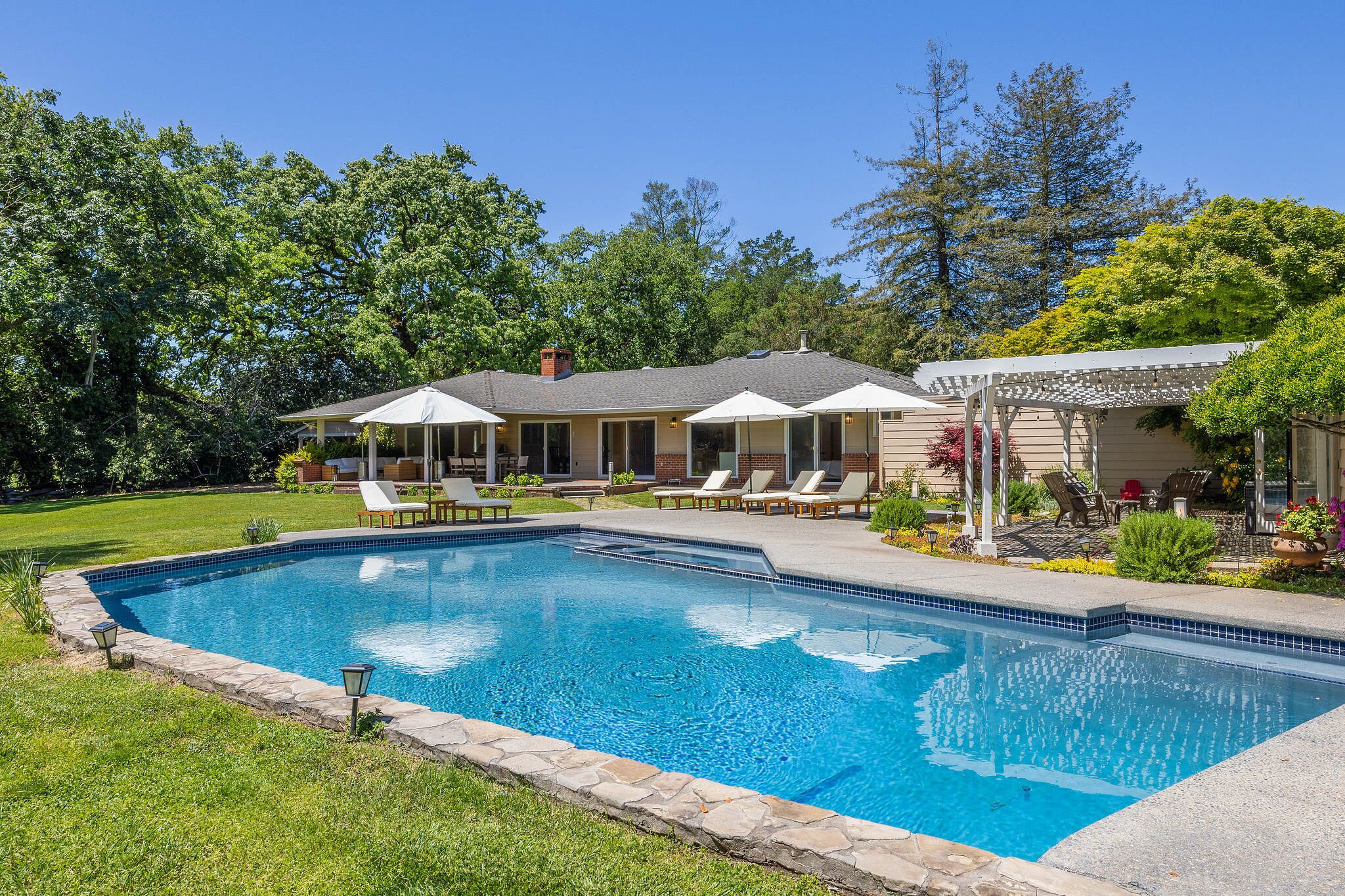 A swimming pool with lounge chairs and umbrellas, surrounded by a well-maintained lawn and trees.