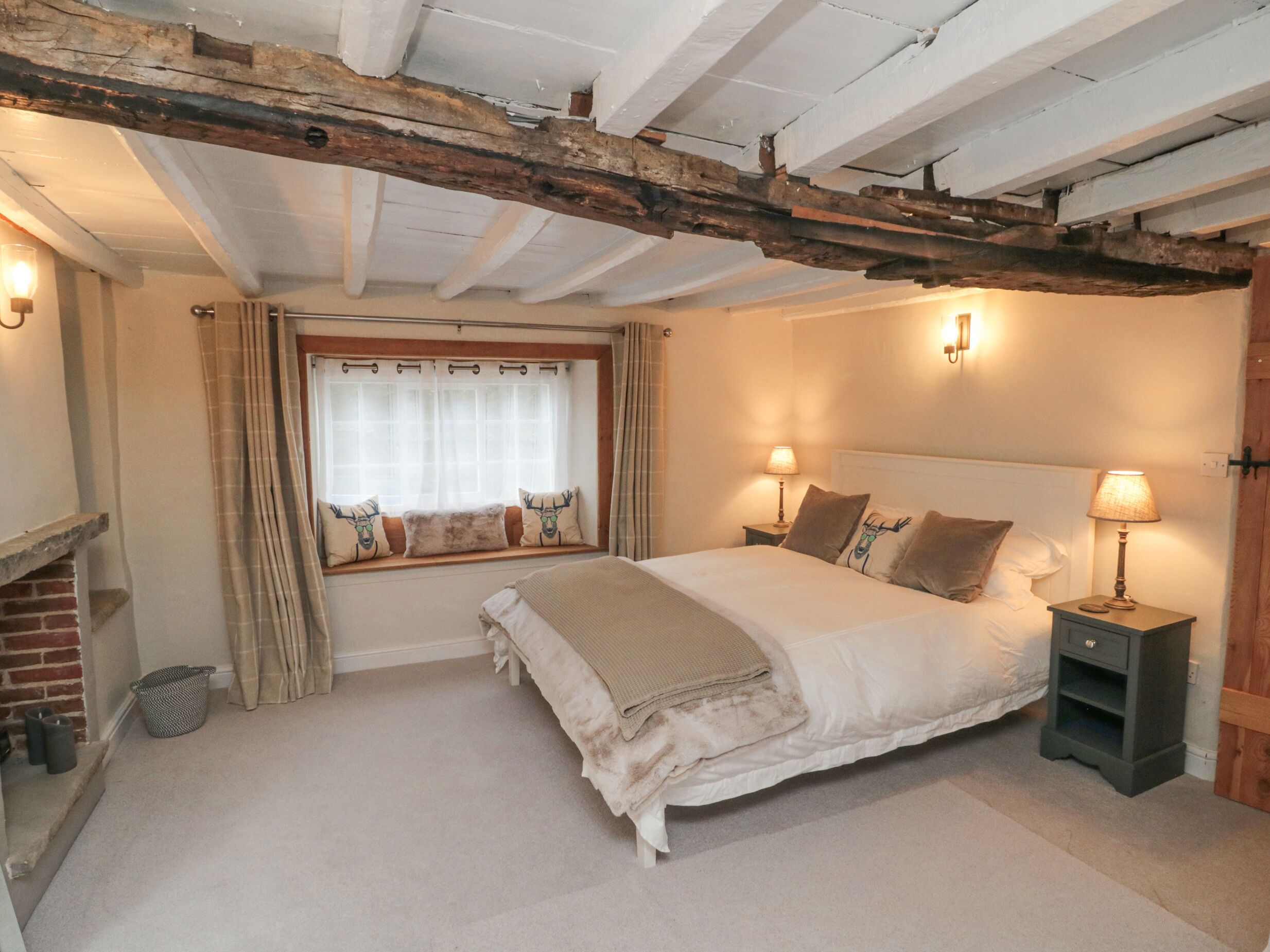 A bedroom with a large bed, a window with curtains, a wooden beam ceiling, and a nightstand with a lamp.