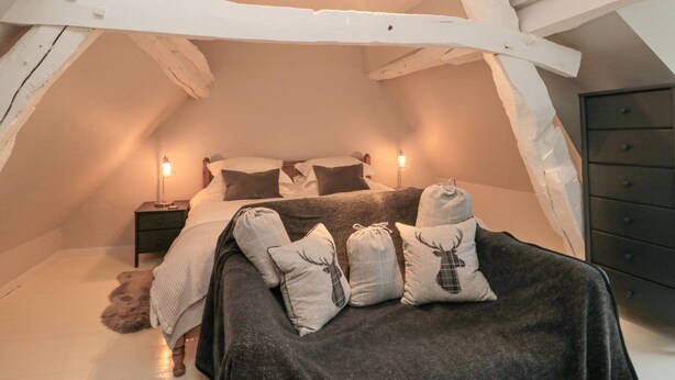 A cozy attic bedroom with exposed wooden beams, a bed with multiple pillows, and a hanging light fixture. - York House (Pickering)