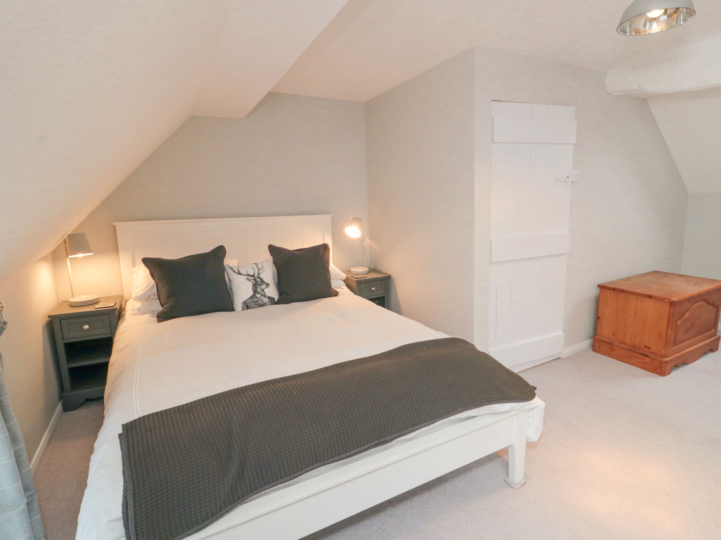 A bedroom with a white bed, dark pillows, a bedside lamp, a nightstand, and a wooden chest.