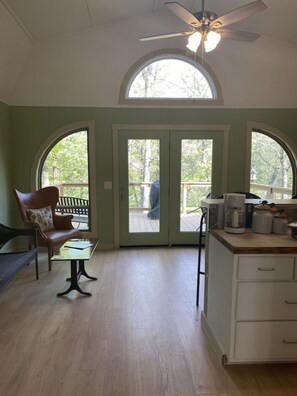 A room with a wooden floor, a ceiling fan, a bench, a table, and a kitchen area with a coffee maker and jars.