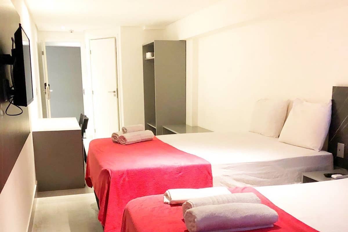 Habitación de hotel con dos camas, cada una con colcha roja y almohadas blancas, y un televisor de pantalla plana montado en la pared.