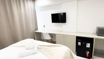 Quarto de hotel moderno com cama, escrivaninha, cadeira, televisão e minibar.