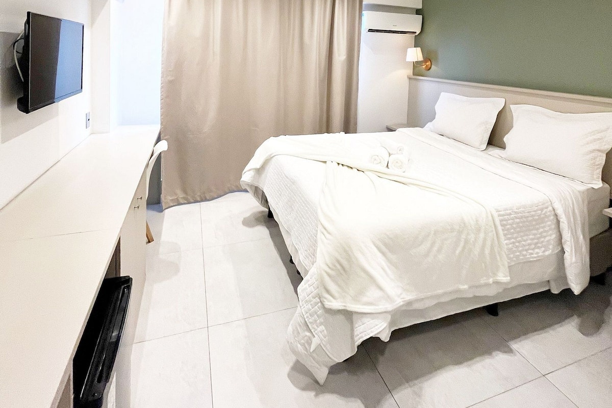 Una habitación de hotel con cama, televisor montado en la pared, escritorio y ventana con cortinas.