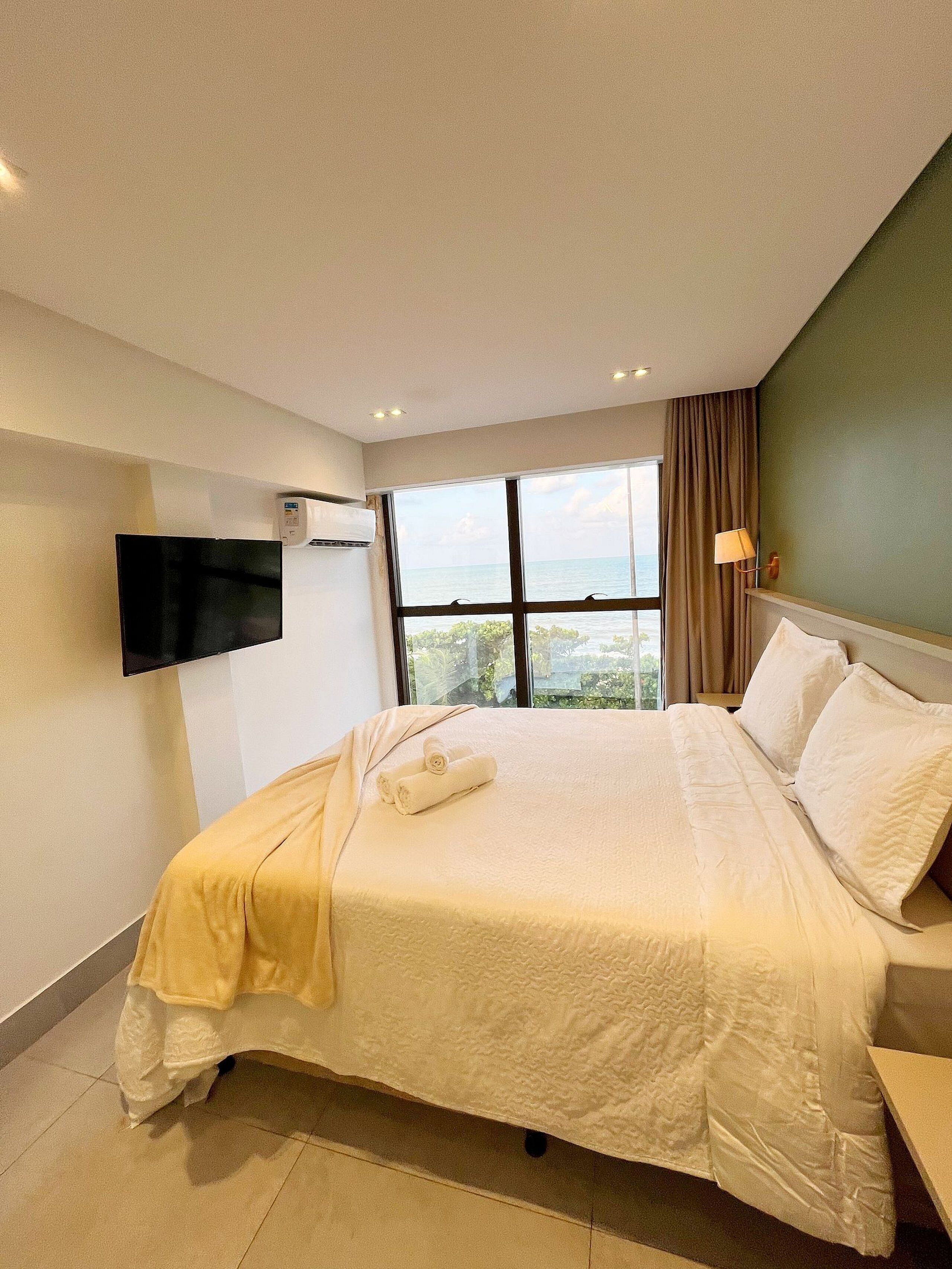 Une chambre d’hôtel avec un grand lit, un téléviseur à écran plat fixé au mur, une fenêtre donnant sur la mer et une lampe sur la table de chevet.