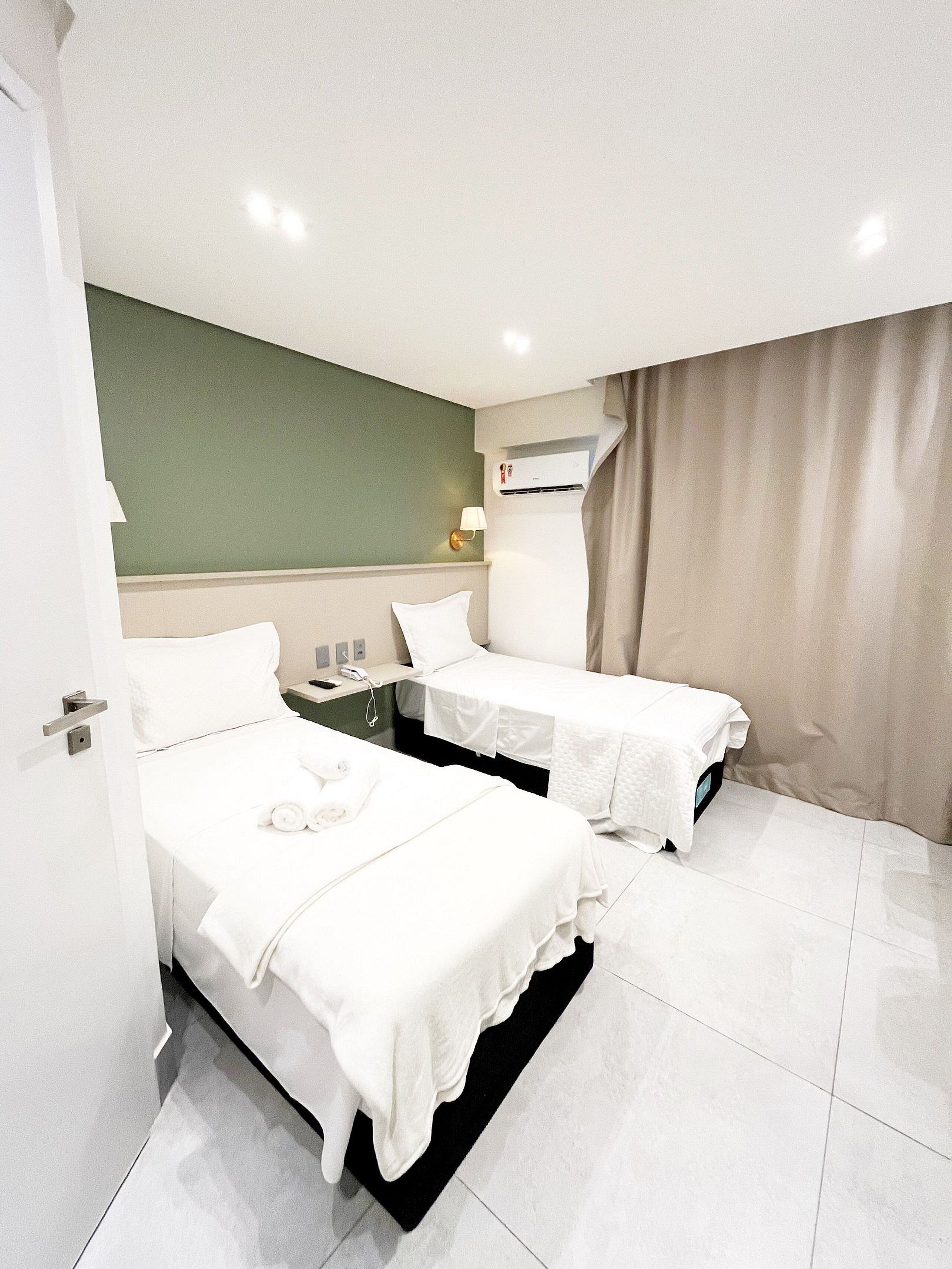 Ein Hotelzimmer mit zwei Betten, einer grünen Wand, einer Klimaanlage, einer Lampe und einem Vorhang.