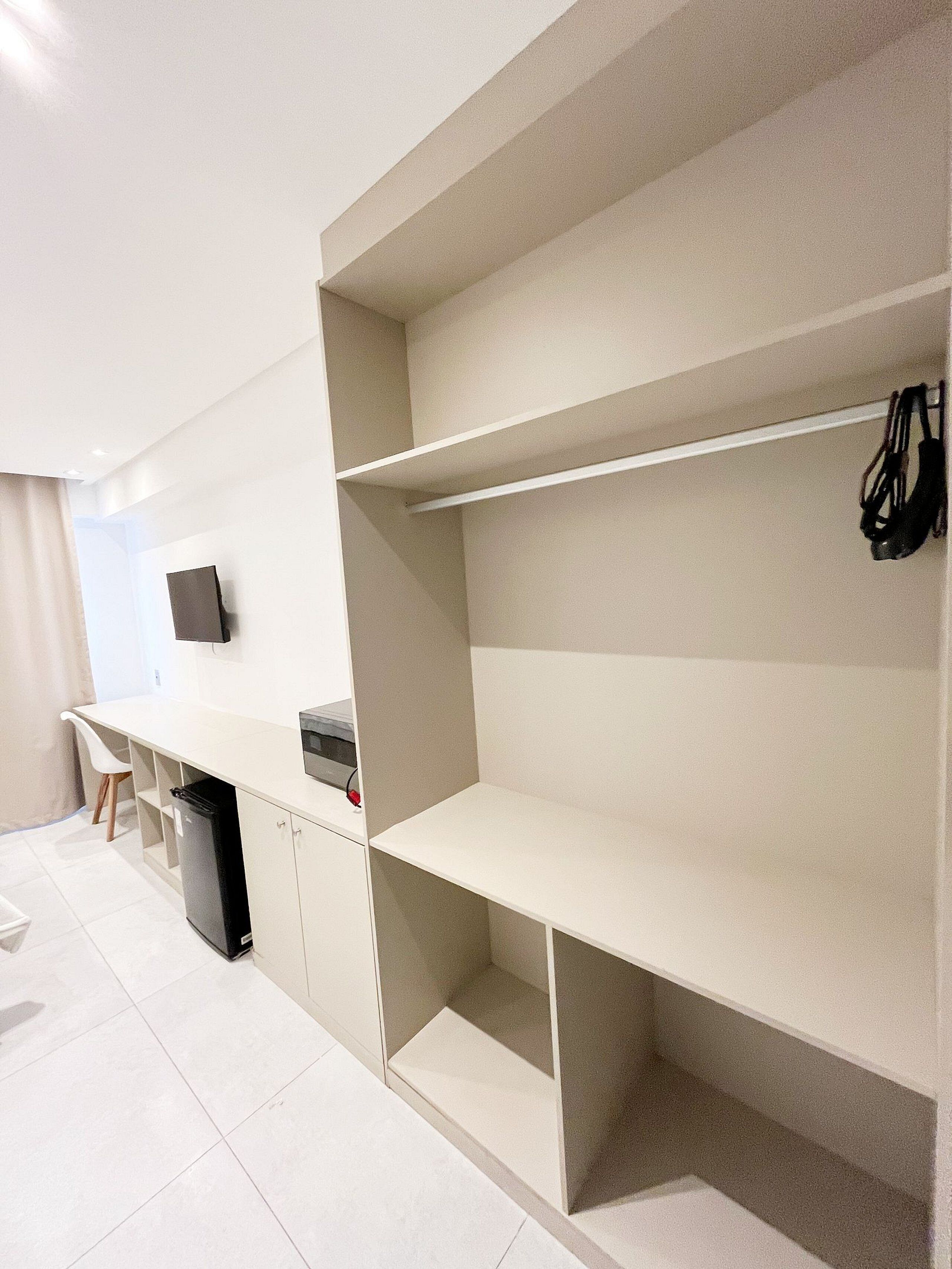 A modern, minimalist room with white walls, a flat-screen TV mounted on the wall, a small desk with a chair, and a black trash bin.