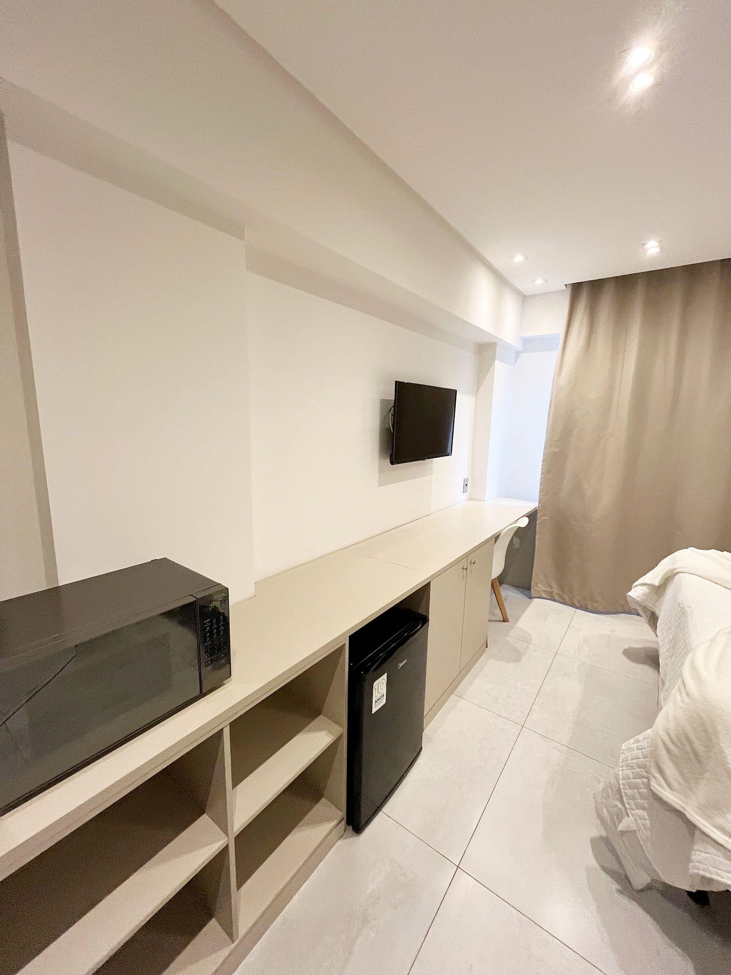 Quarto de hotel com cama, escrivaninha com televisão, micro-ondas e mini-geladeira.