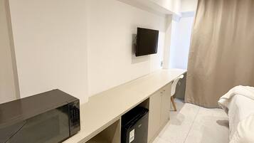 Quarto de hotel com cama, escrivaninha com televisão, micro-ondas e mini-geladeira.