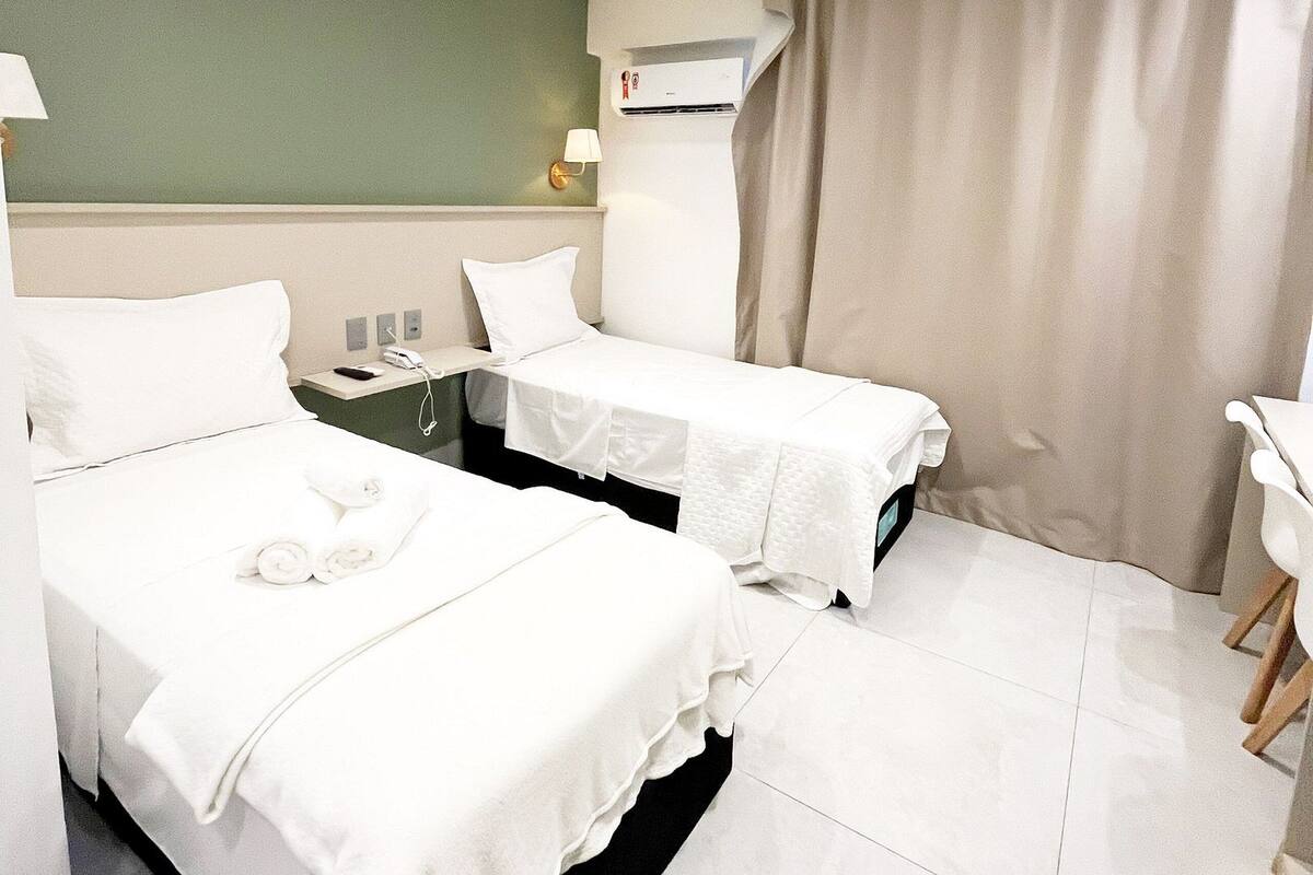 Habitación de hotel con dos camas, una mesita de noche con lámpara, un equipo de aire acondicionado y una cortina.