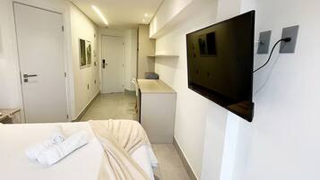 Ein Hotelzimmer mit einem Bett, einem an der Wand befestigten Fernseher, einer kleinen Küchenzeile und einer Tür zu einem weiteren Raum.