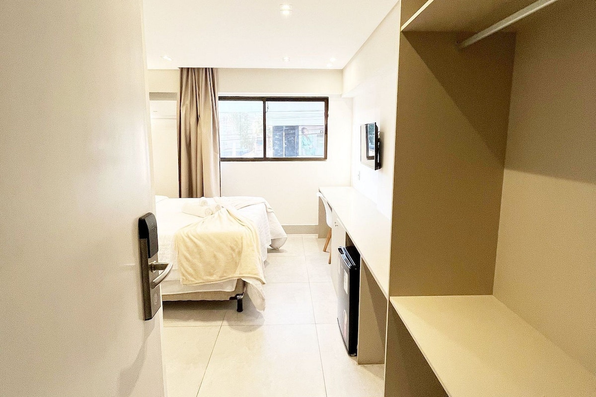 Habitación de hotel con cama, escritorio, silla, ventana y estante empotrado.