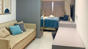 A modern hotel room with a sofa, a bed with a blue bedspread, a desk, and a large mirror. - Arpoar Suites By Day By Day (João Pessoa)
