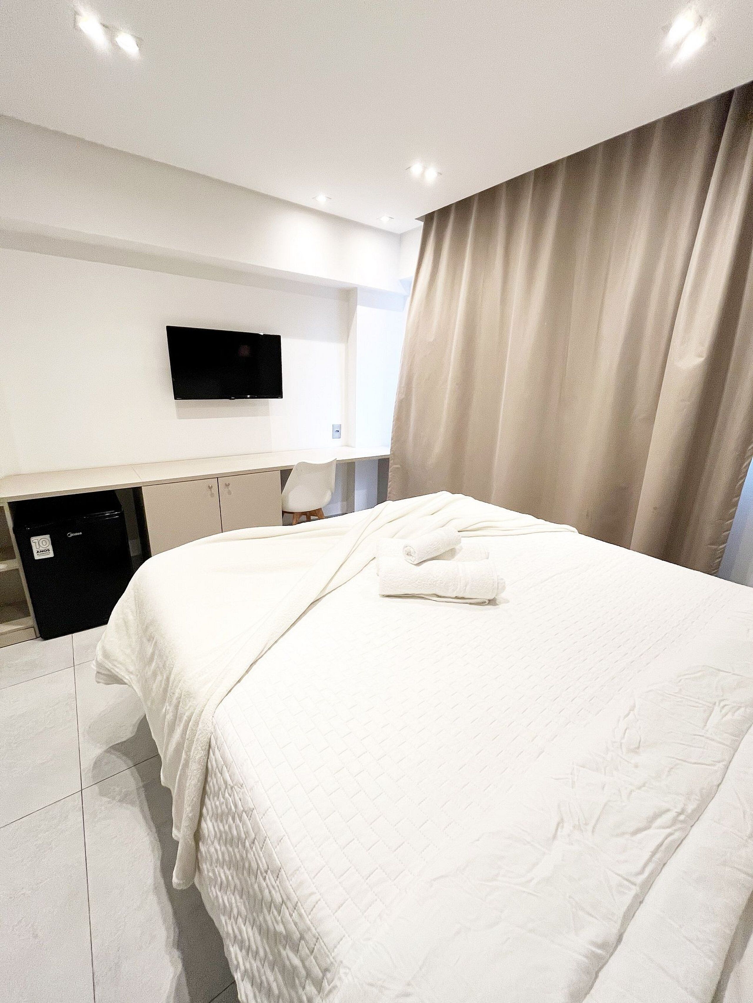 A hotel room with a bed, a TV mounted on the wall, a desk, and a window with curtains.