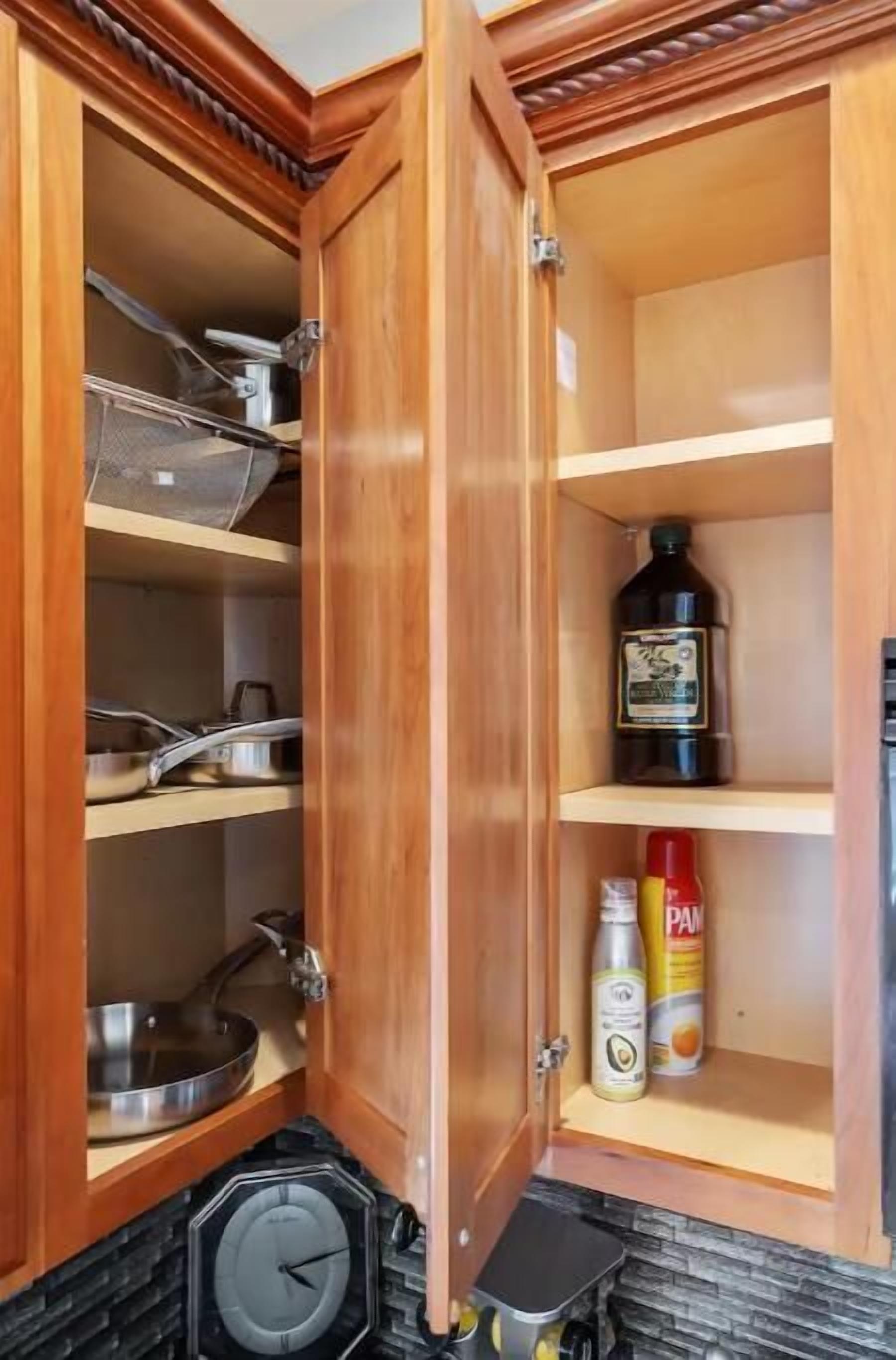 A wooden kitchen cabinet with shelves containing cookware, a bottle of cooking oil, and a can of sauce.