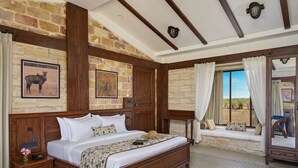 Une chambre à coucher avec un grand lit, une tête de lit en bois, des murs en pierre, un coin lecture près de la fenêtre et une table de chevet avec une lampe.