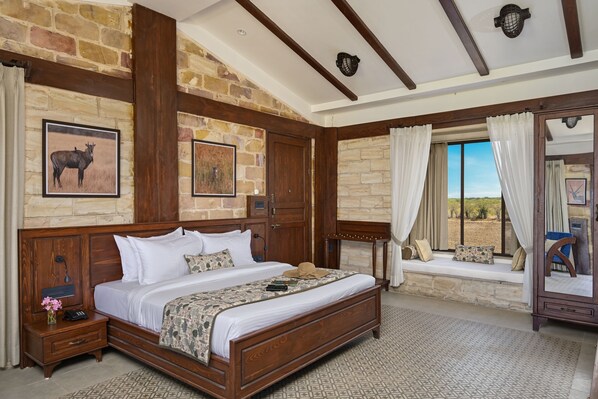 A bedroom with a large bed, wooden headboard, stone walls, a window seat, and a nightstand with a lamp. - The Blackbuck Trails Velavadar (Bhavnagar)