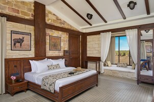 Un dormitorio con una cama grande, cabecera de madera, paredes de piedra, un rincón con asiento junto a la ventana y una mesita de noche con lámpara.