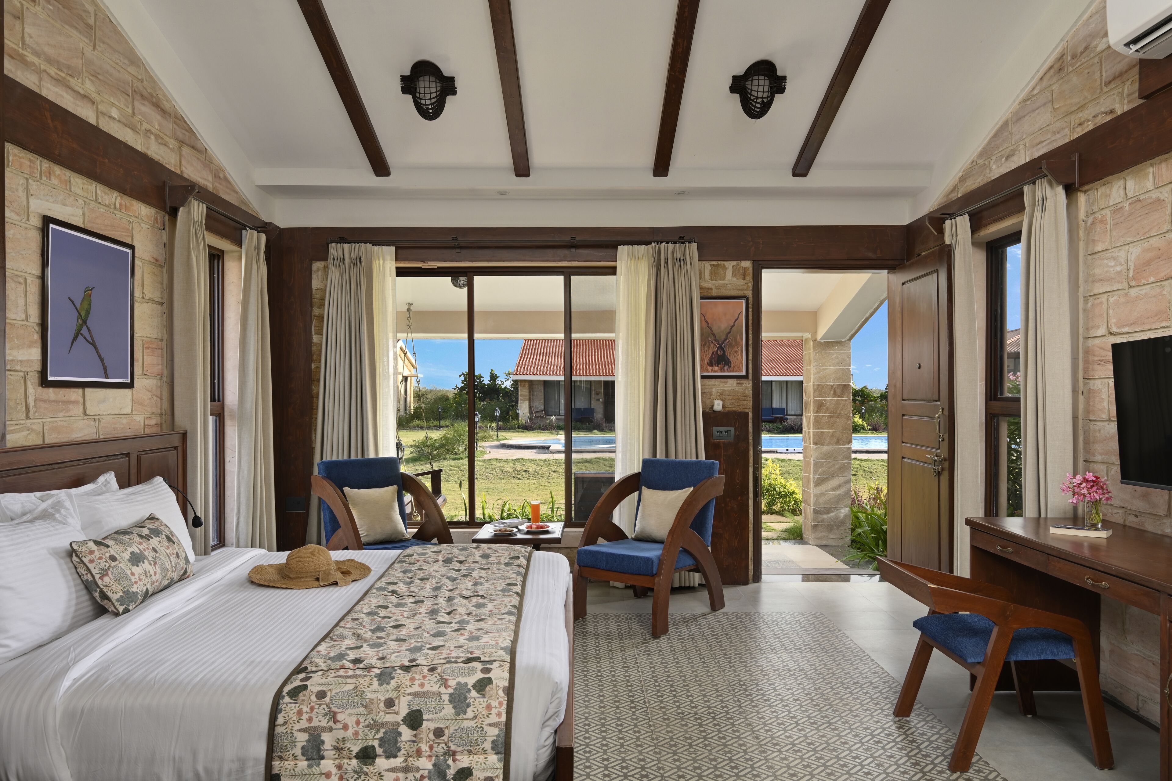A hotel room with a bed, two chairs, a desk, and a large window with a view of a building and a pool.
