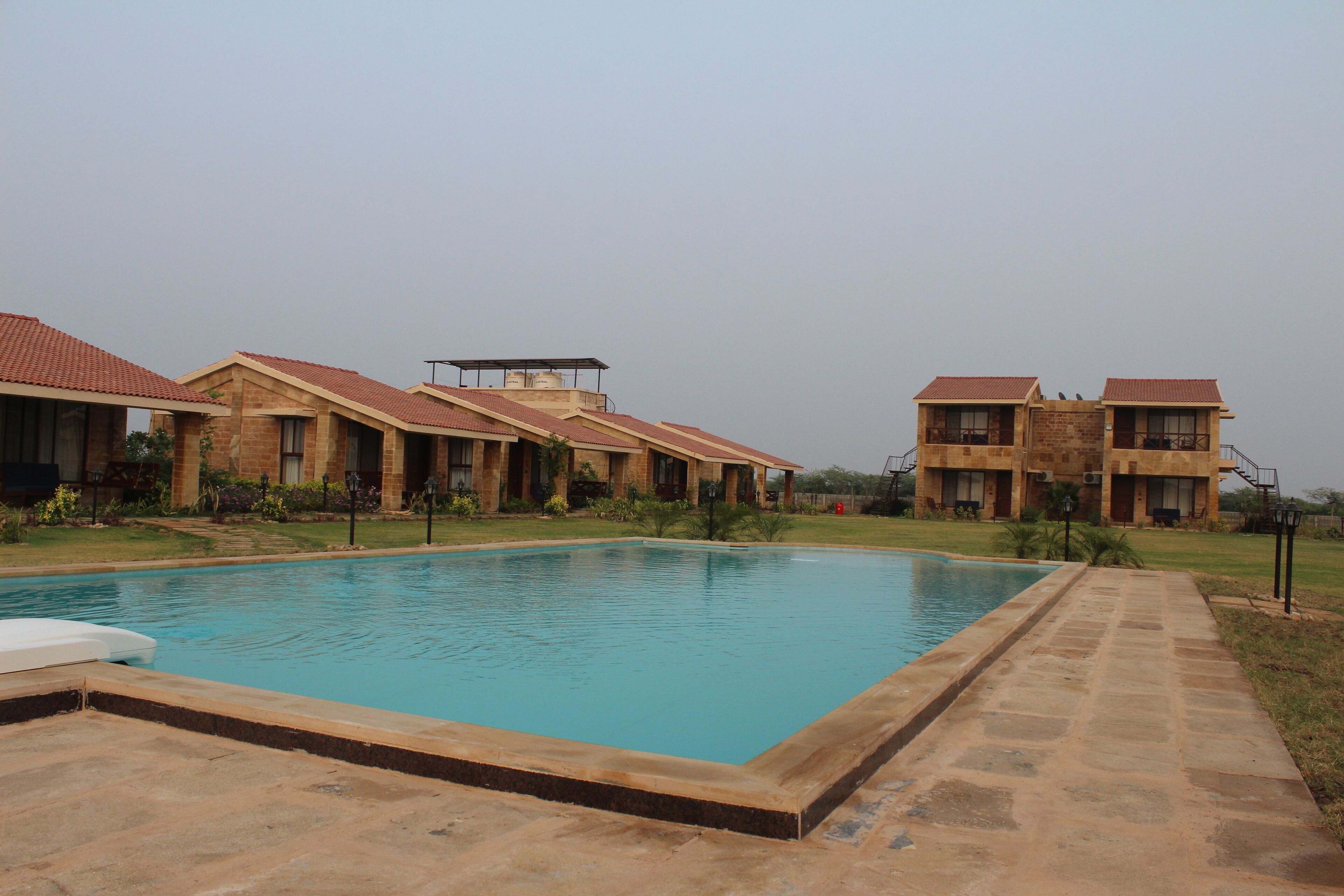 A swimming pool surrounded by brick buildings with red roofs, a clear sky, and a well-maintained lawn.