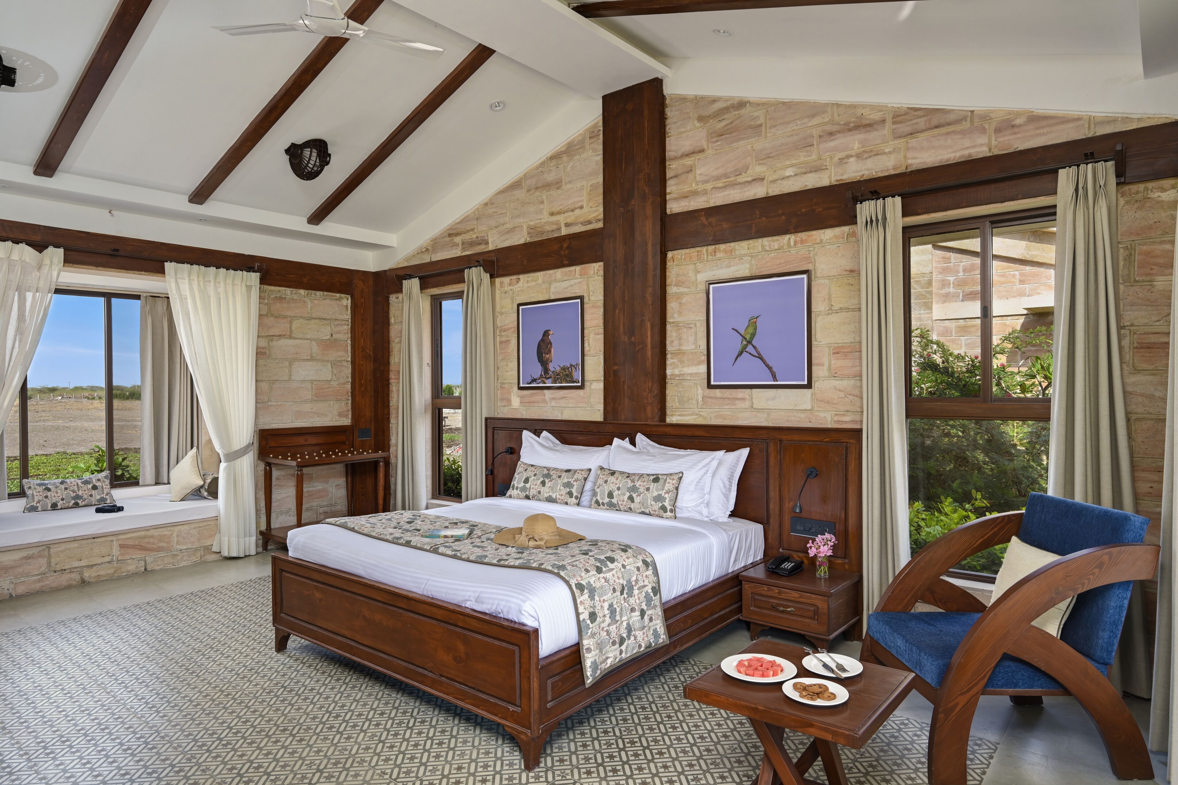 Un dormitorio espacioso con una cama grande, cabecera de madera, mesita de noche, una silla y vistas al exterior a través de ventanas amplias.