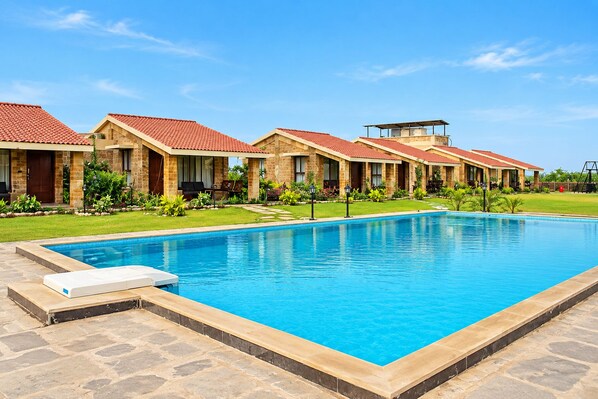 A swimming pool with a stone border, a lounge chair, and a row of buildings with red-tiled roofs. - The Blackbuck Trails Velavadar (Bhavnagar)