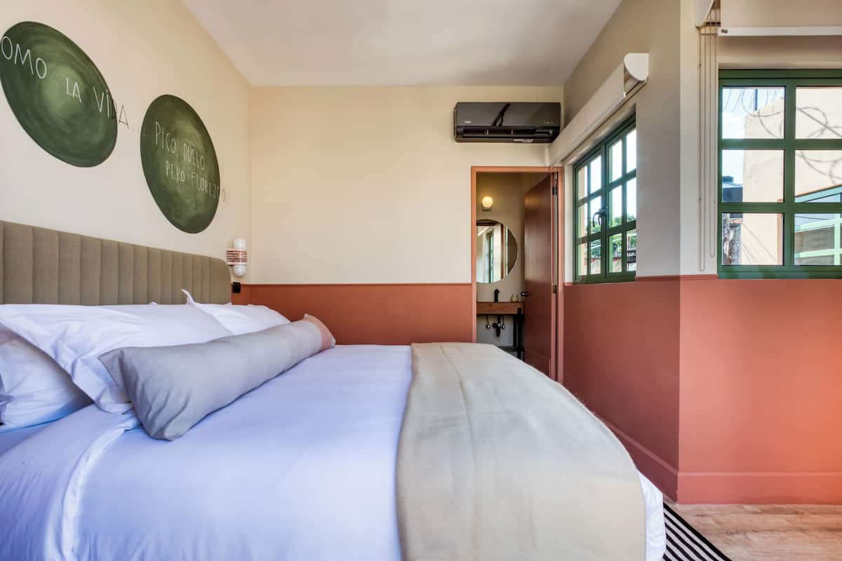 A hotel room with a bed, a wall-mounted air conditioner, and a window with a view.
