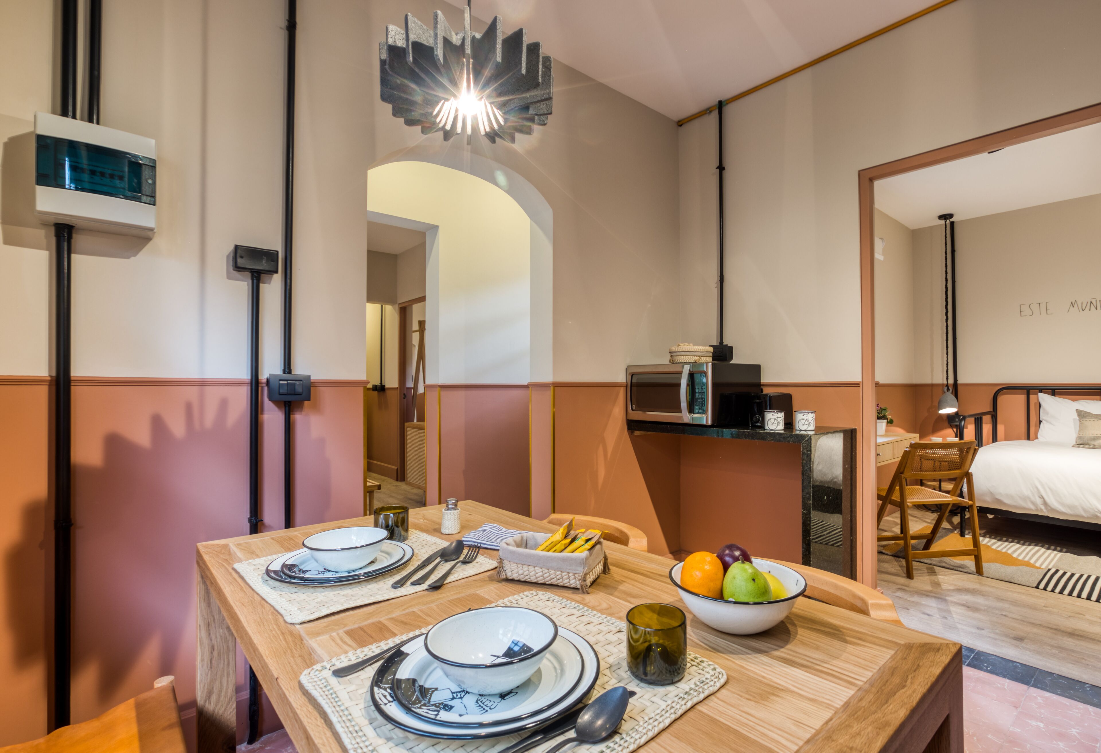 A modern hotel room with a dining area featuring a wooden table set with plates, bowls, and cutlery. A fruit bowl and a small basket of bread are on the table. The room has a bed with a black frame, a bedside table with a lamp, and a wall-mounted air conditioning unit.