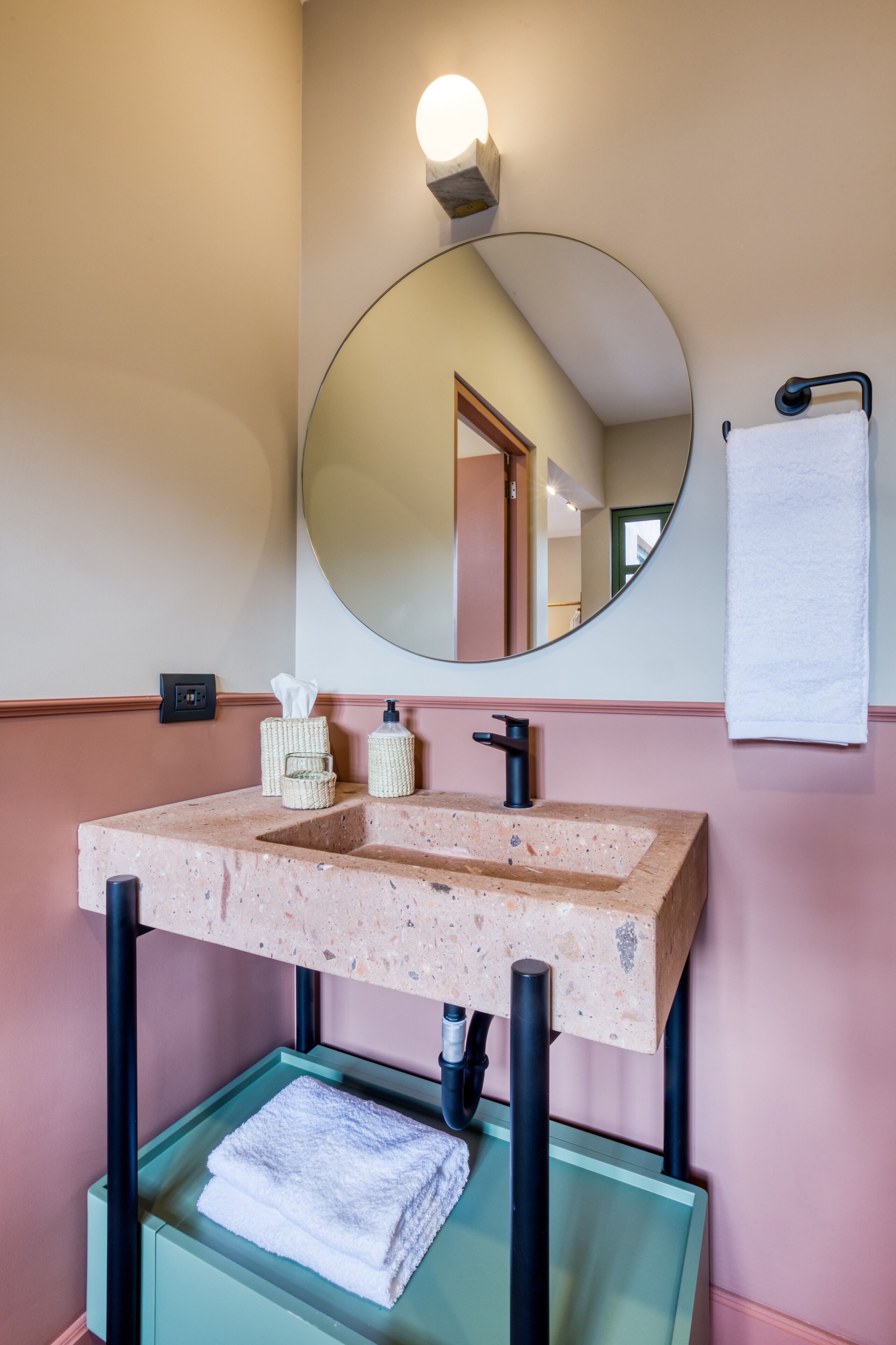 Un baño con lavamanos independiente, un espejo redondo, un perchero y una lámpara de pared.