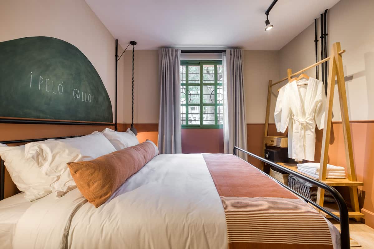 A hotel room with a bed, a chalkboard with text, a window with curtains, and a wooden rack with clothes.