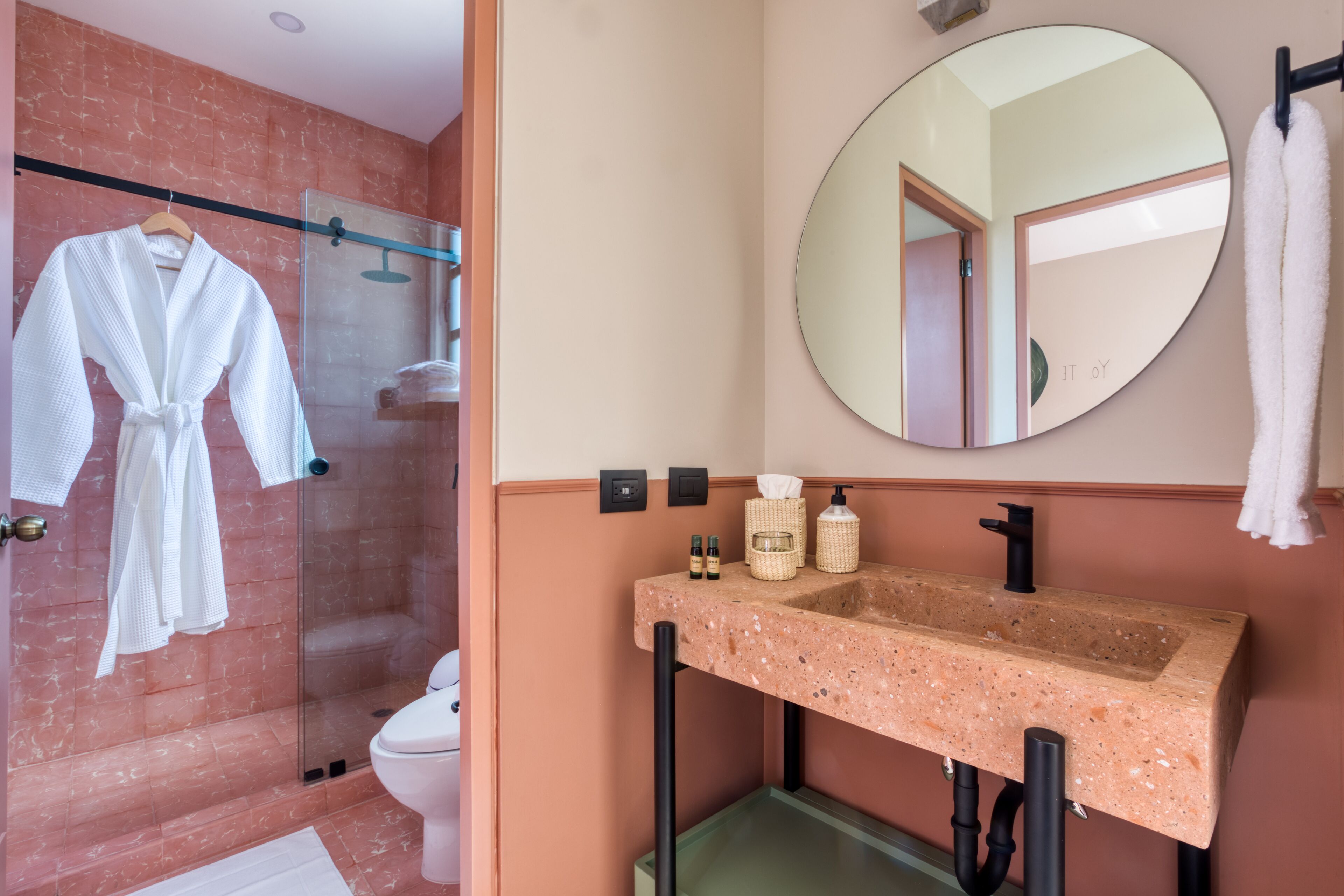 Un baño con cabina de ducha de vidrio, pared con azulejos de mármol, lavabo con encimera salpicada y un espejo redondo, además de un perchero para toallas.