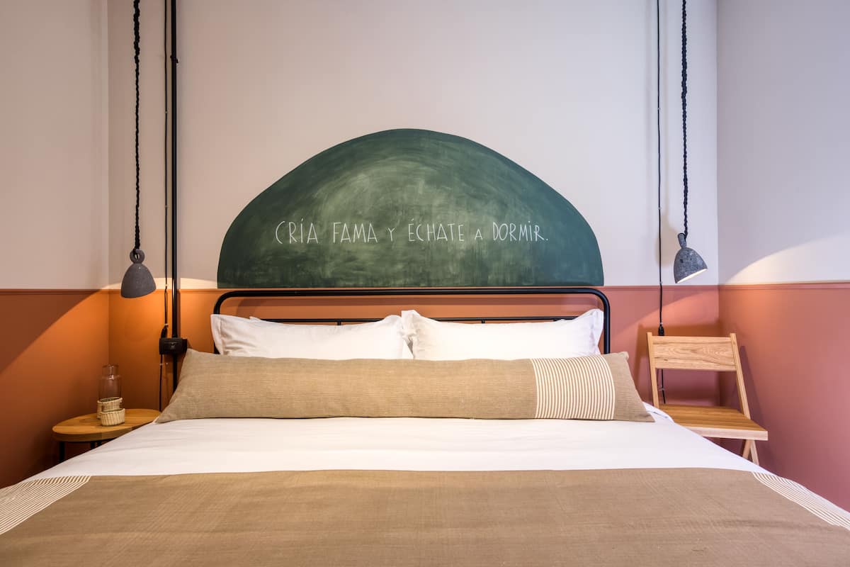 A neatly made bed with a headboard that has a curved design and a motivational quote. There are two bedside tables with lamps, a small wooden chair, and a wall-mounted shelf.